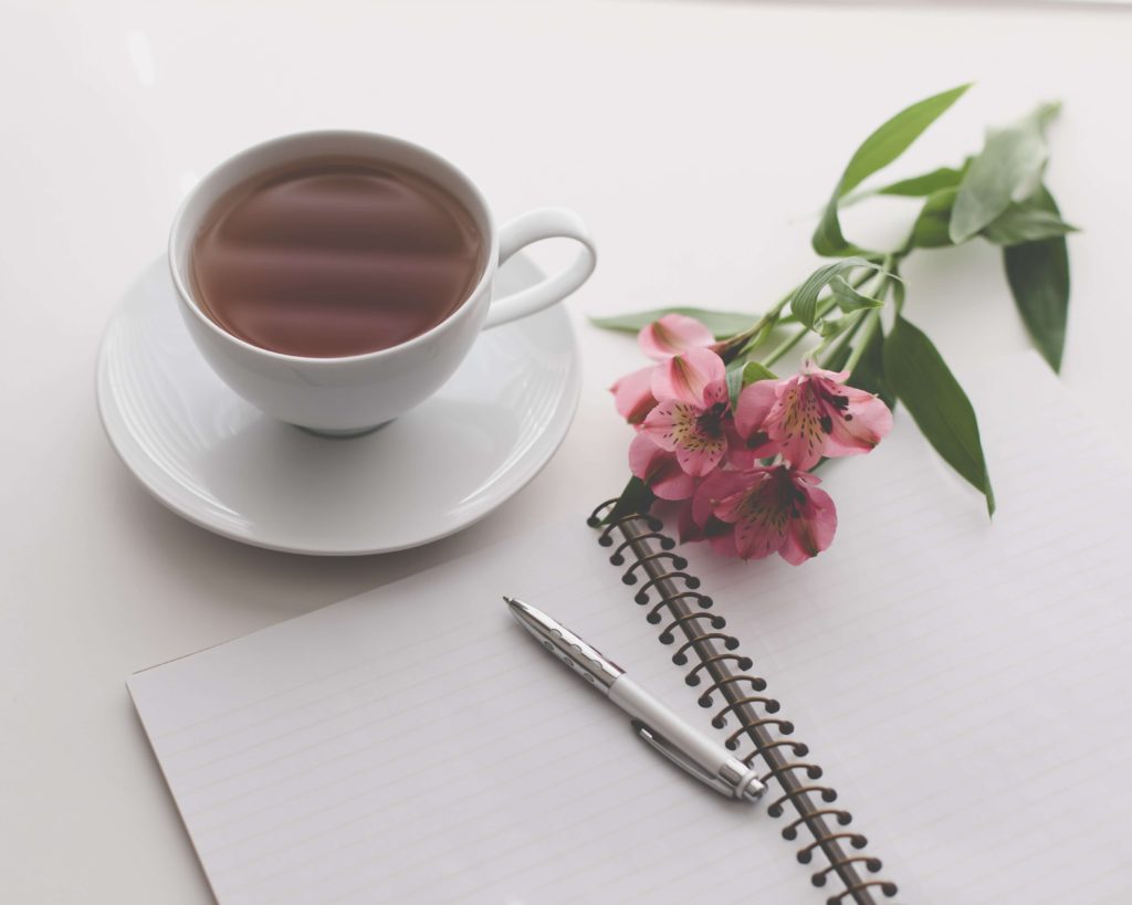 Utiliser les affirmations positives: une tasse de thé posée près d'un brin de fleurs roses sous un cahier vierge avec un stylo.